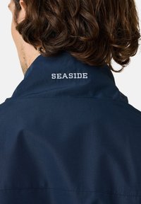 Marinejakke med struktureret stof, krave og det broderede ord "SEASIDE" i hvidt på bagsiden.
