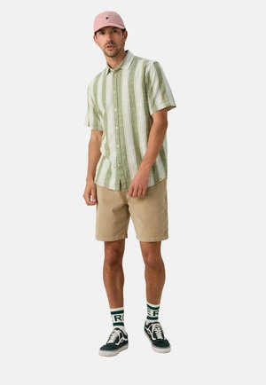 Korte mouwen overhemd met groene en witte verticale strepen, gecombineerd met beige shorts, zwarte sneakers en een roze pet.