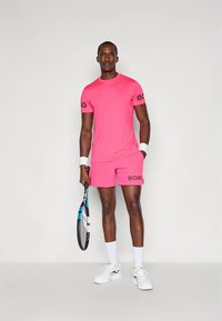Ljusrosa sportoutfit med korta ärmar och shorts med "BORG"-logotyp i svart. Modellen håller en tennisracket och bär vita sneakers.