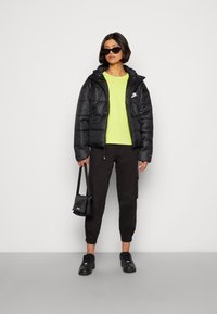 Svart puffjakke med hette og logo, neongrønn t-skjorte, svarte cargo-bukser og svarte joggesko. Modellen holder en liten svart sekk.