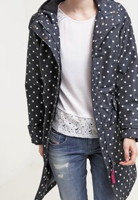 Impermeabile blu marino a pois con zip frontale, grande cappuccio e bottoni a pressione. Indossato sopra una maglietta bianca con bordi in pizzo e jeans strappati.