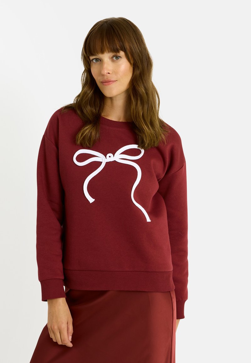 Sudadera color burdeos con un diseño de lazo blanco en el centro. Cuello redondo y puños acanalados. Textura suave, combinada con una falda borgoña brillante.