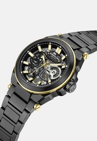 Automatyczny zegarek z czarnym stalowym paskiem, czarnymi i złotymi akcentami, szkieletowym cyferblatem odsłaniającym mechanizmy oraz mocnymi znacznikami godzinowymi.