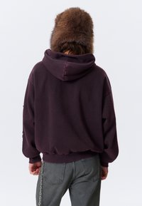 Person iført en mørk lilla hættetrøje, grå jeans med en kædeaccessory og en stor brun pelsagtig hat, der vender væk fra kameraet.