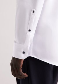 Witte dressshirt met een eenvoudige structuur, voorzien van een hexagonale manchet en marineblauwe knopen. Een geborduurd zwart roosdetail bevindt zich op de manchet.