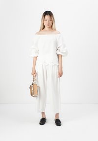 Top bianco scollo off-the-shoulder con maniche a palloncino, ricamo floreale all'orlo, abbinato a pantaloni bianchi a gamba larga. Scarpe nere e una piccola borsa beige.