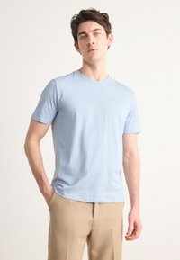 T-shirt in cotone azzurro chiaro con collo rotondo e maniche corte. Presenta un piccolo logo bianco sul lato sinistro del petto. Abbinato a pantaloni beige.