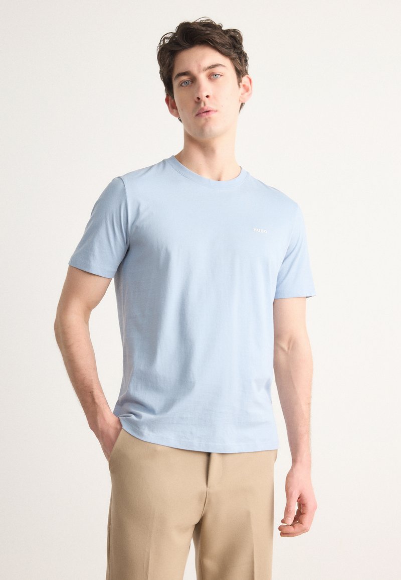 T-shirt in cotone azzurro chiaro con collo rotondo e maniche corte. Presenta un piccolo logo bianco sul lato sinistro del petto. Abbinato a pantaloni beige.
