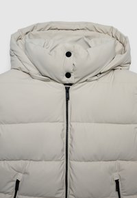 Giacca imbottita beige con colletto alto, cappuccio staccabile, zip nera, bottoni a pressione e tasche laterali con zip.
