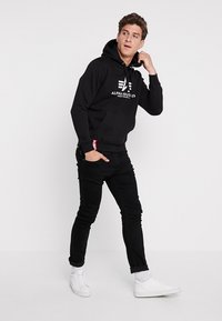 Zwarte hoodie met een logo aan de voorkant, een buidelzak en verstelbare koordjes; gedragen met zwarte jeans en witte sneakers.