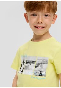 Lichtgele katoenen T-shirt met korte mouwen, met een fotoprint van golven en surfplanken. De tekst luidt "OCEAN is mijn speeltuin."