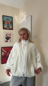 Balta plīsētā jaka ar augstu apkakli un priekšējo rāvējslēdzēju. Raksturīgas vaļīgas piedurknes un gluda, spīdīga tekstūra.