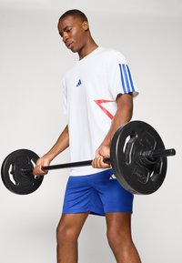Weißes Sportshirt mit blauen Streifen und roten Akzenten, kombiniert mit blauen Shorts. Person hebt eine schwarze Langhantel mit Gewichten.