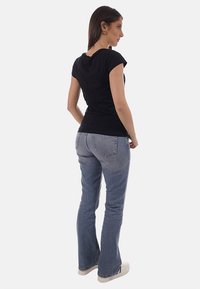 Donna in piedi di profilo, che indossa una maglietta aderente nera a maniche corte, jeans a zampa di elefante azzurri chiaro e scarpe da ginnastica bianche su uno sfondo semplice.