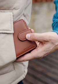Brown leather wallet with a snap closure is partially pulled from a beige puffer jacket pocket, showcasing smooth texture and embossed brand logo.