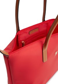 Κόκκινη τσάντα ώμου με καφέ λαβές, μερικώς ανοιχτό φερμουάρ που αποκαλύπτει εσωτερική τσέπη και μικρή δερμάτινη ετικέτα με την επωνυμία στο εσωτερικό.