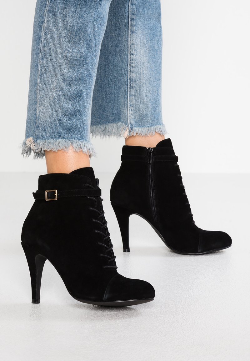 Bottines noires en daim à talons hauts avec fermeture éclair latérale, laçage avant et bride à boucle portées avec un jean bleu à ourlet effiloché.