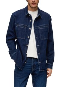 Donkerblauwe denim blouse met een knoopsluiting aan de voorkant, twee borstzakken en witte stiksels. Model draagt het over een witte shirt.