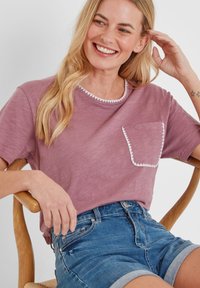 Smiling woman with long blonde hair wearing a mauve t-shirt with white trim on pocket and collar, seated on a wooden chair in denim shorts.