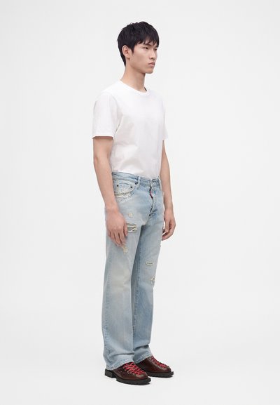 Jeune homme debout portant un t-shirt blanc uni, un jean bleu clair déchiré et des chaussures de randonnée marron avec des lacets rouges.