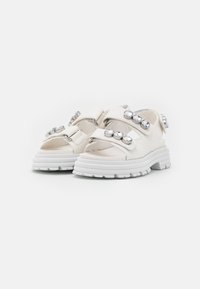 Sandales blanches avec une épaisse semelle blanche, plusieurs sangles en Velcro et des accents décoratifs en argent sur les sangles. Texture en cuir lisse.