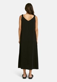 Schwarzes ärmelloses Kleid mit V-Ausschnitt im Rücken, das versammelte Träger und eine lockere Passform hat, die bis unter die Knöchel reicht. Glattes Gewebe.