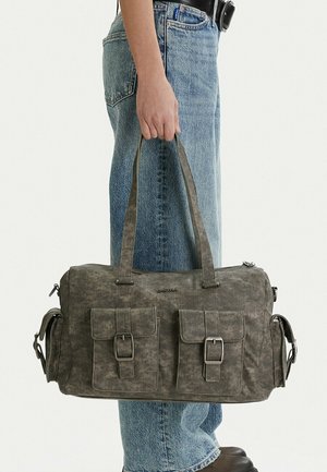 Persona sosteniendo una bolsa de lona gris con dos bolsillos delanteros con hebillas, de pie con vaqueros azules y zapatos negros sobre fondo blanco.