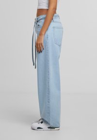 Osoba oblečená v světle modrých širokých džínách a bílých teniskách, stojící bokem proti jednoduchému šedému pozadí.