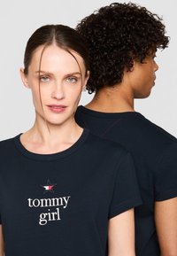 Tamsiai mėlynos trumpomis rankovėmis marškinėliai; priekinėje pusėje yra „tommy girl“ tekstas ir žvaigždutės logotipas; gale matomas raudonas siuvinėjimo detalė.