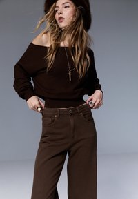 Maglione marrone con scollo a barca e orlo a costine, abbinato a jeans a gamba larga a vita alta di colore marrone scuro. Dettagli in oro di grande effetto.