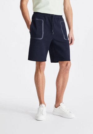 Uomo con pantaloncini neri al ginocchio con tasche anteriori delineate, camicia bianca e sneaker bianche, in piedi su sfondo bianco.