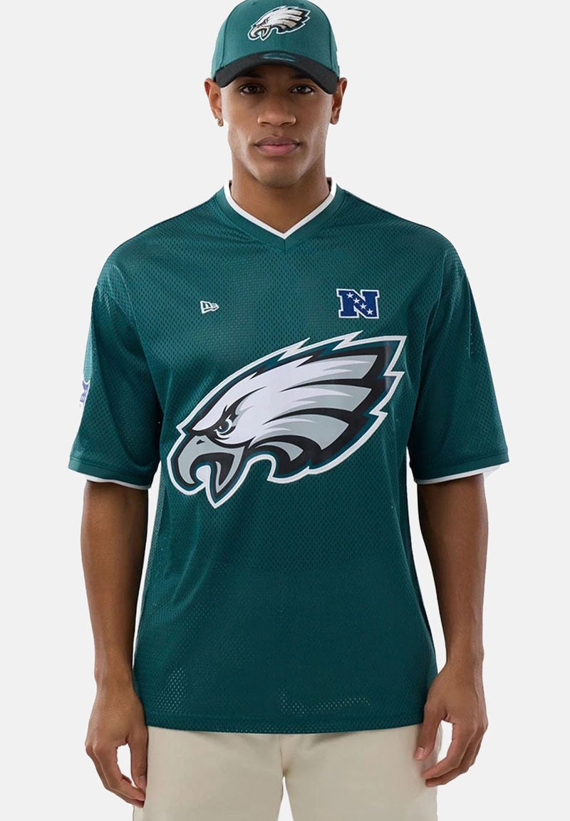 Homme portant un maillot NFL des Philadelphia Eagles vert foncé et une casquette assortie, debout devant un fond blanc uni.