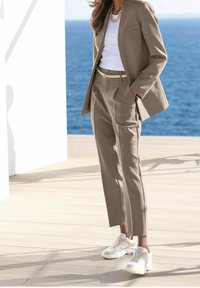 Kobieta w beżowym garniturze z dopasowanymi spodniami, białym paskiem, białej bluzce i masywnych białych sneakersach, stoi na drewnianym tarasie przy wodzie.