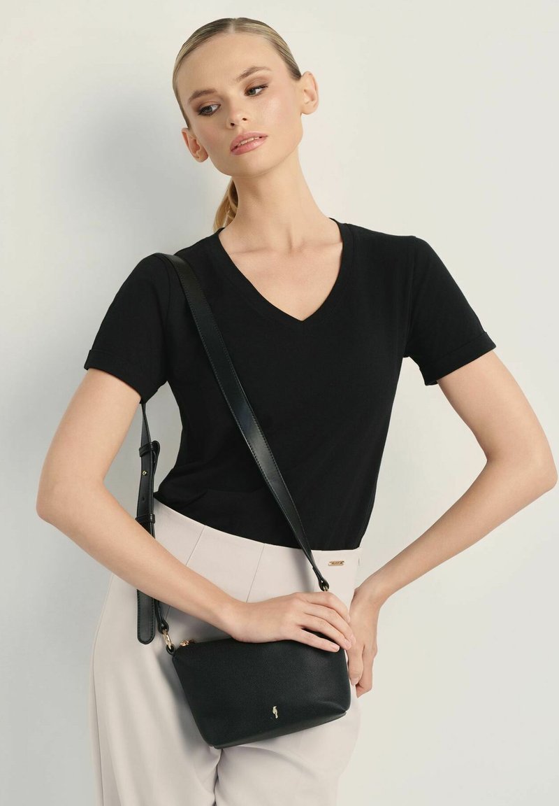 Czarna skórzana torba na ramię z złotym akcentem, prostokątny kształt i regulowany pasek, w zestawieniu z czarnym T-shirtem z dekoltem w serek i jasnymi spodniami.