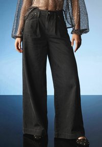 Korkeavyötäröiset, leveälahkeiset mustat farkut, joissa on pleatit, viisi taskua ja metallinen napitus. Yhdistettynä läpikuultavaan tekstuuriin.