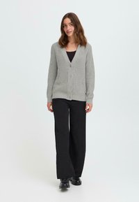 Cardigan côtelé gris clair avec un col en V, fermeture à boutons et manches longues, associé à un pantalon large noir et des bottes noires.