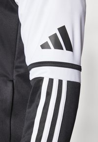 Giacca atletica bianca e nera con tre strisce nere sulla manica e logo Adidas nero sulla parte superiore del braccio; tessuto liscio e leggero.