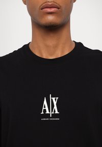 Juodas medvilninis marškinėliai su apvali apykakle, centre esantis balintas siuvinėjimo "A|X" logotipas ir tekstas "ARMANI EXCHANGE".