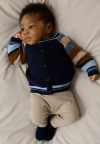 Knitted cardigan in navy, with multicolored striped sleeves; paired with beige pants and navy socks, resting on soft, light-colored fabric.