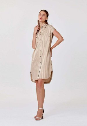 Femme portant une robe boutonnée à rayures verticales beige et blanche, à manches courtes, et des talons blancs à bride à la cheville, debout devant un fond uni.