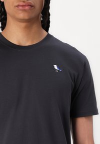 Schwarzes Baumwoll-T-Shirt mit kurzen Ärmeln, mit einer kleinen weißen und blauen Vogelstickerei auf der linken Brustseite. Sanfte Textur.