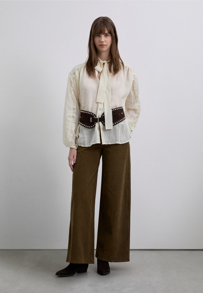 Blouse claire texturée avec un col à nouer, associée à un pantalon large en velours côtelé marron et une ceinture en cuir foncé ornée de clous métalliques.