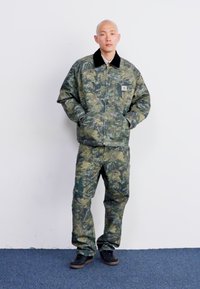 Set giacca e pantaloni mimetici con texture trapuntata in tonalità di verde e marrone. La giacca presenta un colletto nero e una tasca frontale.