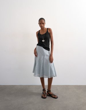 Femme portant un débardeur noir, une jupe évasée rayée jusqu'aux genoux, des sandales noires à lacets et un collier pendentif rond, debout sur un sol gris.