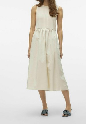 Femme portant une robe midi crème sans manches avec un corsage texturé et une jupe froncée, associée à des sandales plates bleues avec détail fleur en tissu.
