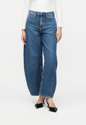 Kvinde iført højtaljede, vidde blå jeans, hvid langærmet top og sorte spidse hæle med spændedetalje.