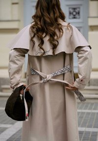 Žena s dlouhými kudrnatými vlasy, oblečená v béžovém trenčkotě, držící černou tkanou tašku a zavazující vzorovaný pásek trenčkotu vzadu.