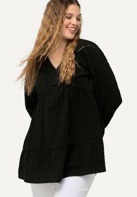 Zwarte, lichte tuniek met een V-hals, lange mouwen en een gelaagde zoom. Beschikt over een textuurstof en decoratieve stiksels langs de halslijn.