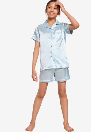 Ung pige står barfodet iført lyseblå satin kortærmet knap-op skjorte og matchende shorts, smilende med hånden på hovedet.