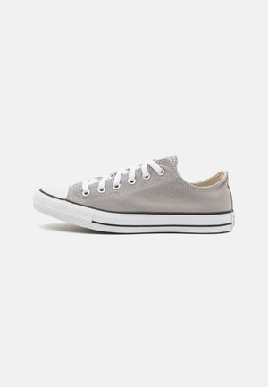 Zapatilla de lona gris con cordones blancos, puntera de goma y franja negra a lo largo de la suela. Diseño de caña baja, textura suave y forma redondeada.